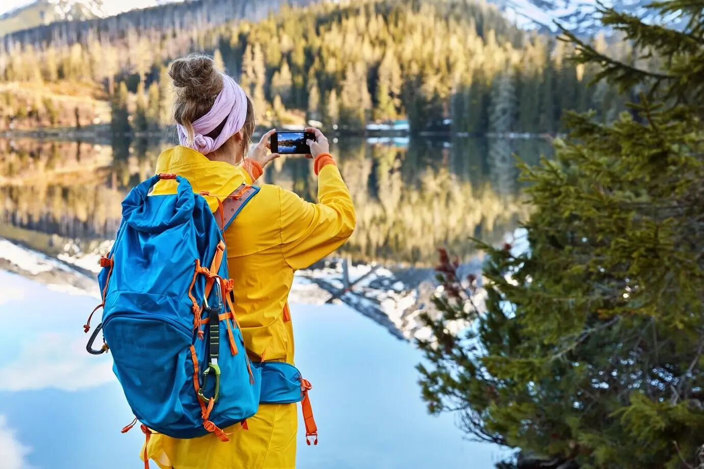 Rear view of an active female tourist using her smartphone to photograph a lakescape with mountains.